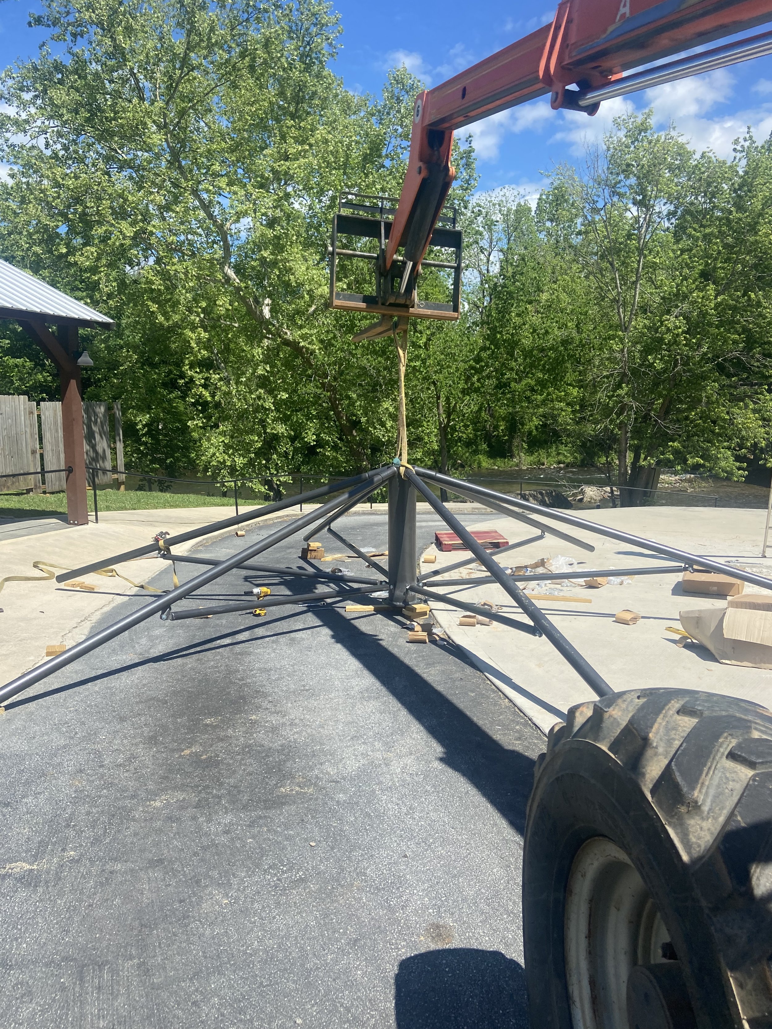 A construction site with a large lift, which is connected to a black metal base with multiple support arms, on a paved surface under a blue sky with scattered clouds and green trees in the background.
