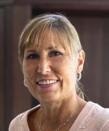 Close-up portrait of a smiling middle-aged woman with blonde hair and earrings, wearing a light pink top.