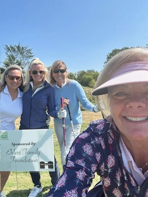 Four women smiling at a golf course, photo taken by one of them, who is wearing a cap and sunglasses. There is a sign that says 'Sponsored by Ehrhart Family Foundation'.