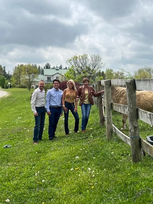 Four people are standing on a grassy field near a wooden fence with a horse or pony grazing. There are houses and trees in the background under a cloudy sky.