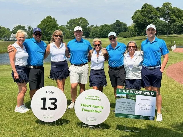 Group of eight people dressed in golf attire standing outdoors on a golf course, holding signs and a large check for a golf tournament winnings, with trees and a clear sky in the background.