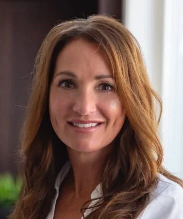 A woman with long, wavy brown hair smiling indoors with a window in the background.
