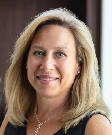 A smiling blonde woman with shoulder-length hair wearing a black sleeveless top and a necklace, indoors with a blurred background.