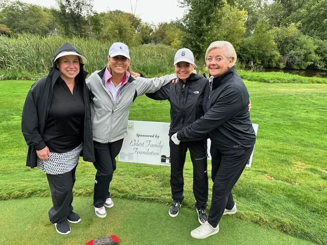 Four women standing together on a golf course, smiling, with trees and a sponsored sign in the background.