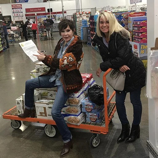 Two women pose with a shopping cart filled with groceries in a store. The woman sitting on the cart is holding a piece of paper, and both are smiling.