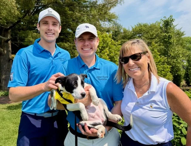 Three people, two young men and an older woman, smiling outdoors holding a black and white puppy with a yellow bandana, surrounded by green trees and grass.