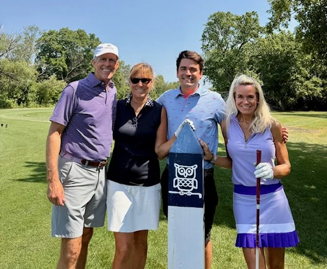 Four people standing outdoors on a golf course, smiling for the camera. They include a man in a purple polo and cap, a woman in a black sleeveless top and white skirt, a man in a light blue patterned shirt, and a woman in a purple and white golf outf