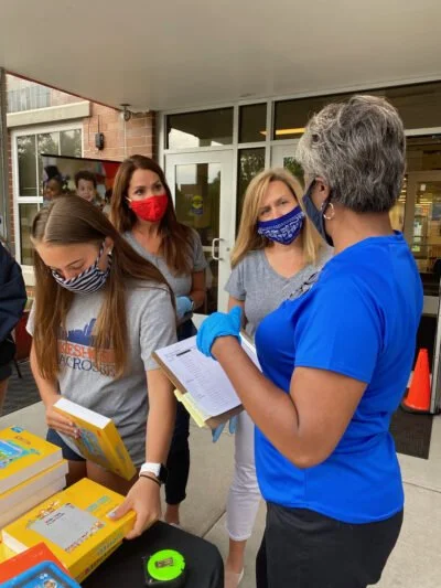 Four women, three of whom are wearing masks, are talking outside a building. One woman in scrubs is holding a clipboard and appears to be explaining something to the others, who are looking at books and materials on a table.