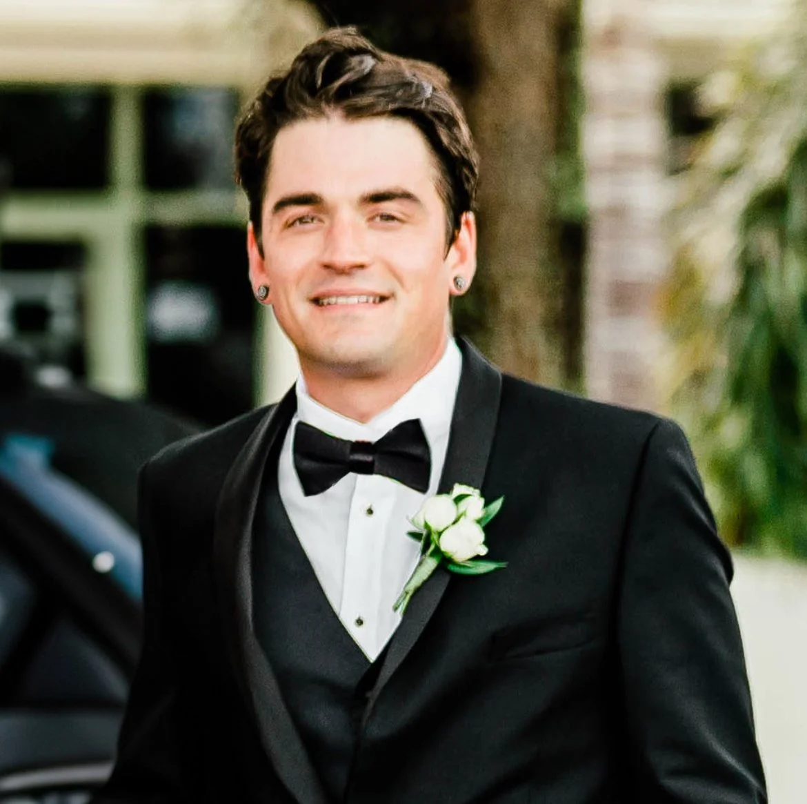 A man dressed in a black tuxedo with a bow tie and a white shirt, smiling at the camera, standing outdoors with greenery in the background.