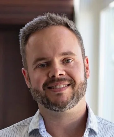A smiling man with short hair and a beard, wearing a light-colored collared shirt, standing indoors near a window.