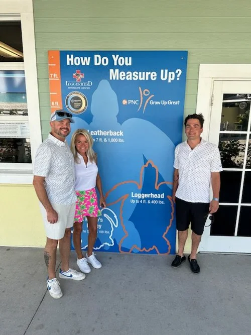 Three people standing in front of a blue height measurement chart with the text 'How Do You Measure Up?' on it. The chart compares a leatherback turtle, measuring up to 7 feet 4 inches and 1,800 pounds, and a loggerhead turtle, which measures up to 4