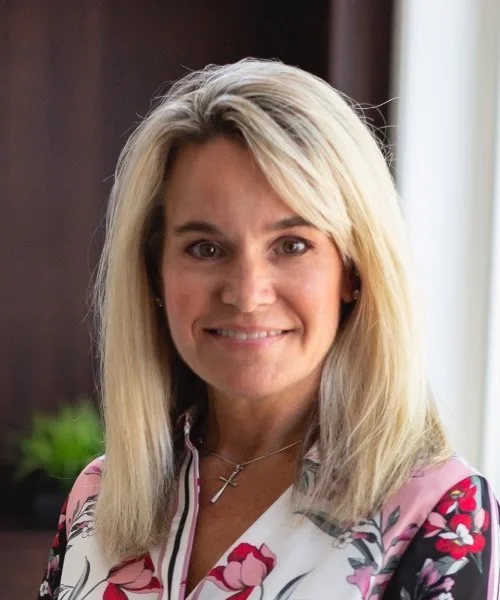 A smiling blonde woman wearing a floral blouse and a cross necklace, standing indoors near a window.