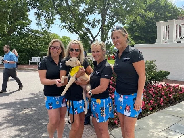 Four women in black shirts and blue shorts with dog-themed patterns stand outdoors under trees, smiling. One woman holds a yellow lab puppy wearing a yellow bandana. People are walking in the background.