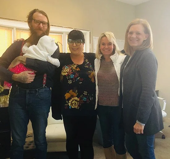 Four adults standing together indoors, with one holding a newborn baby wrapped in a blanket.