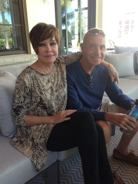 A woman and a man sitting on a white couch in a sunlit room with large windows and tropical plants outside. The woman is wearing a patterned blouse, and the man is in a blue shirt with sunglasses on his head.
