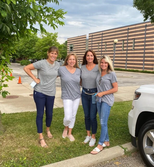 Four women standing outdoors on a grassy area, smiling at the camera, with trees, a building, and a parking lot in the background.