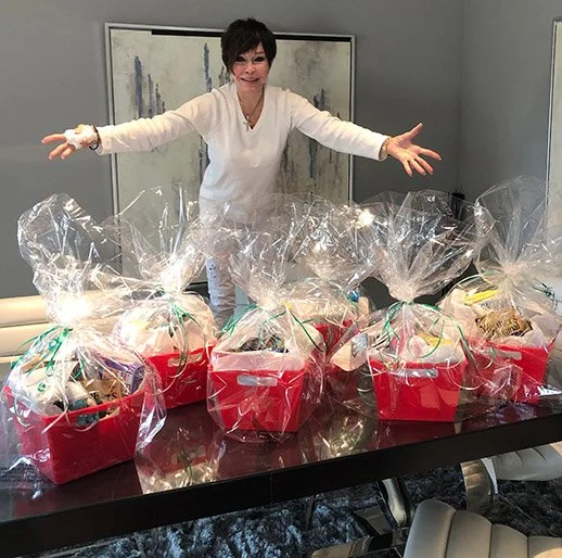 Woman in white sweater smiling with arms outstretched behind a table with multiple wrapped gift baskets in red containers and clear cellophane.