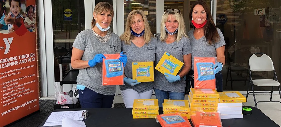 Four women wearing gray shirts and blue gloves stand behind a table with stacks of child ID kits, holding some of the kits and smiling at the camera. One woman has a red face mask around her neck. There is a YMCA banner on the left side of the image