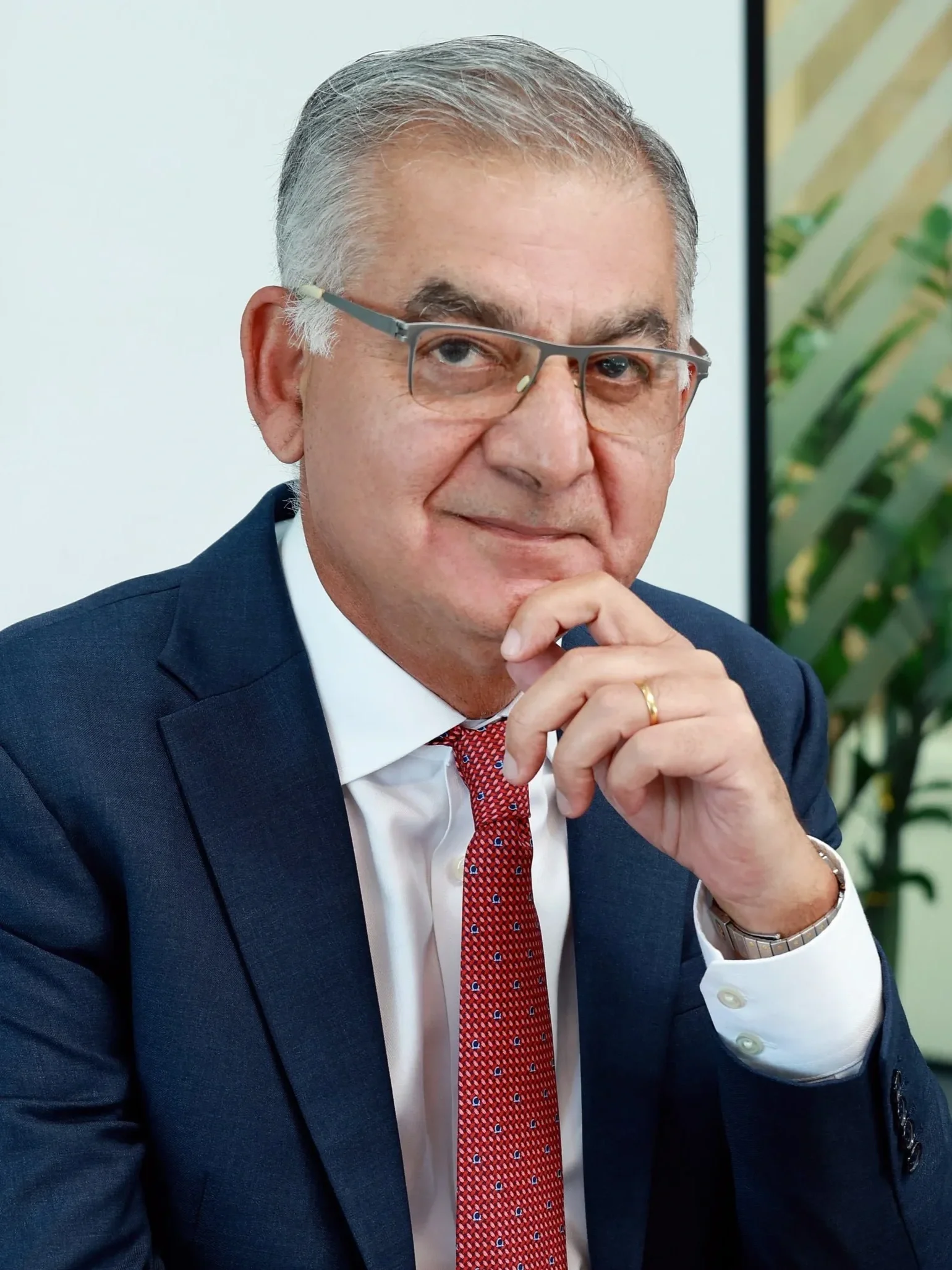 A smiling middle-aged man with slicked-back hair wearing a blue checkered blazer over a light blue shirt, standing outside near a building.