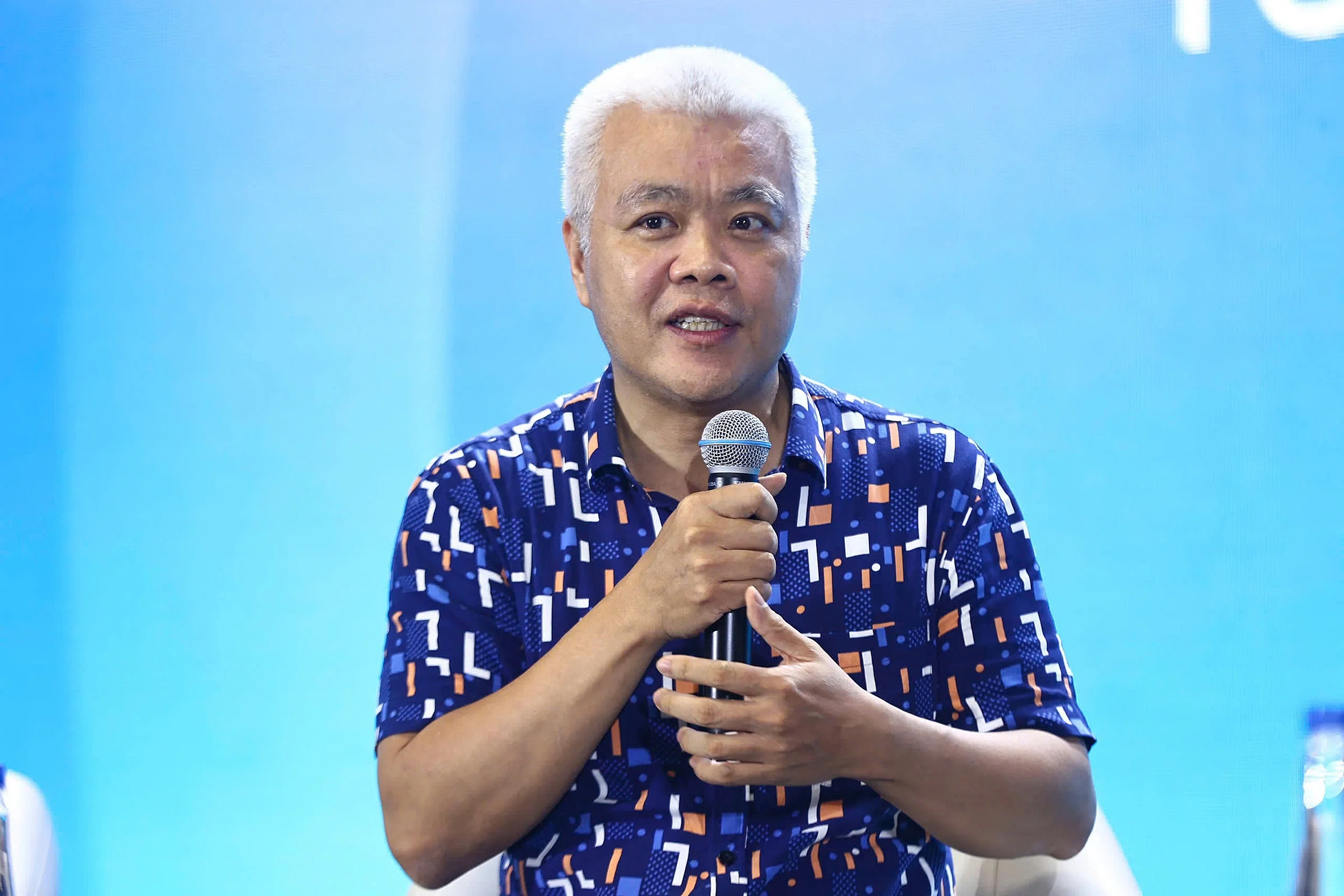 A man with short dark hair, wearing a black polo shirt, standing indoors next to a pink structure, with a plant in the background.