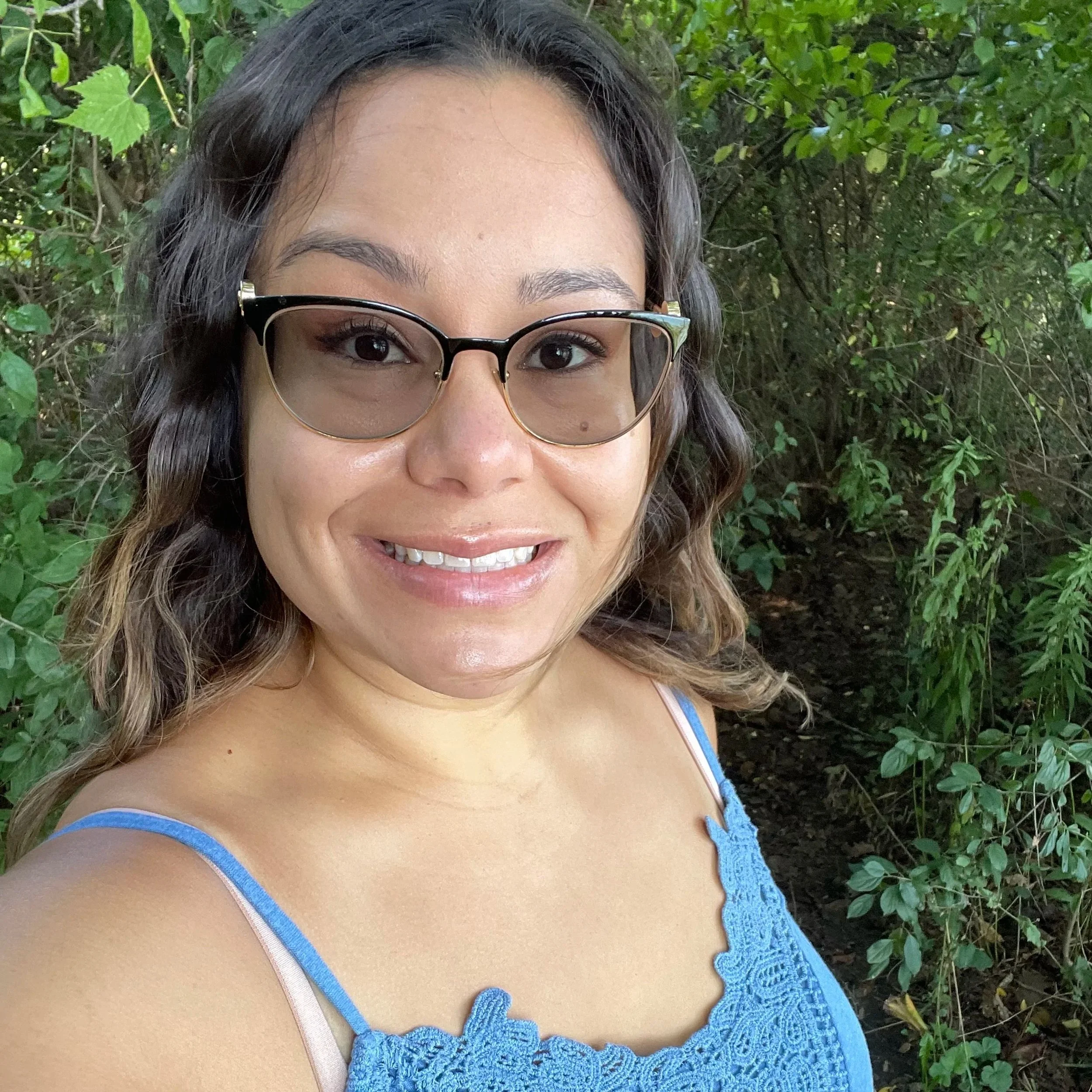 A woman with wavy brown hair, wearing glasses and a blue lace top, smiling outdoors surrounded by green foliage.
