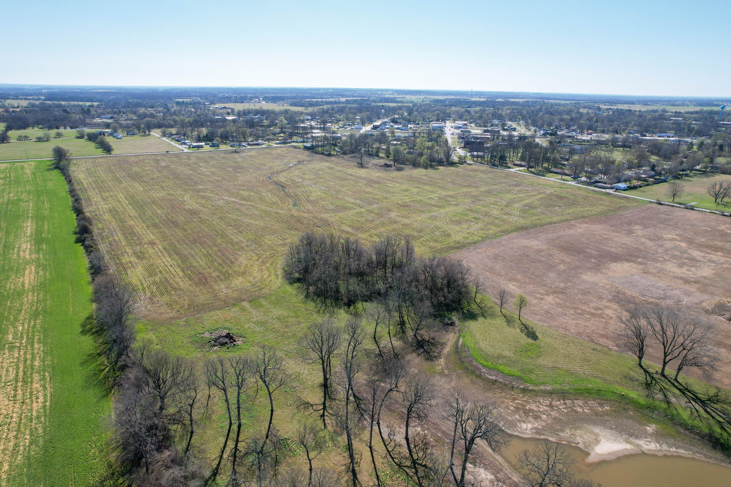 Aerial view of a rural area with open fields, scattered trees, and a small town in the background, under a clear blue sky.