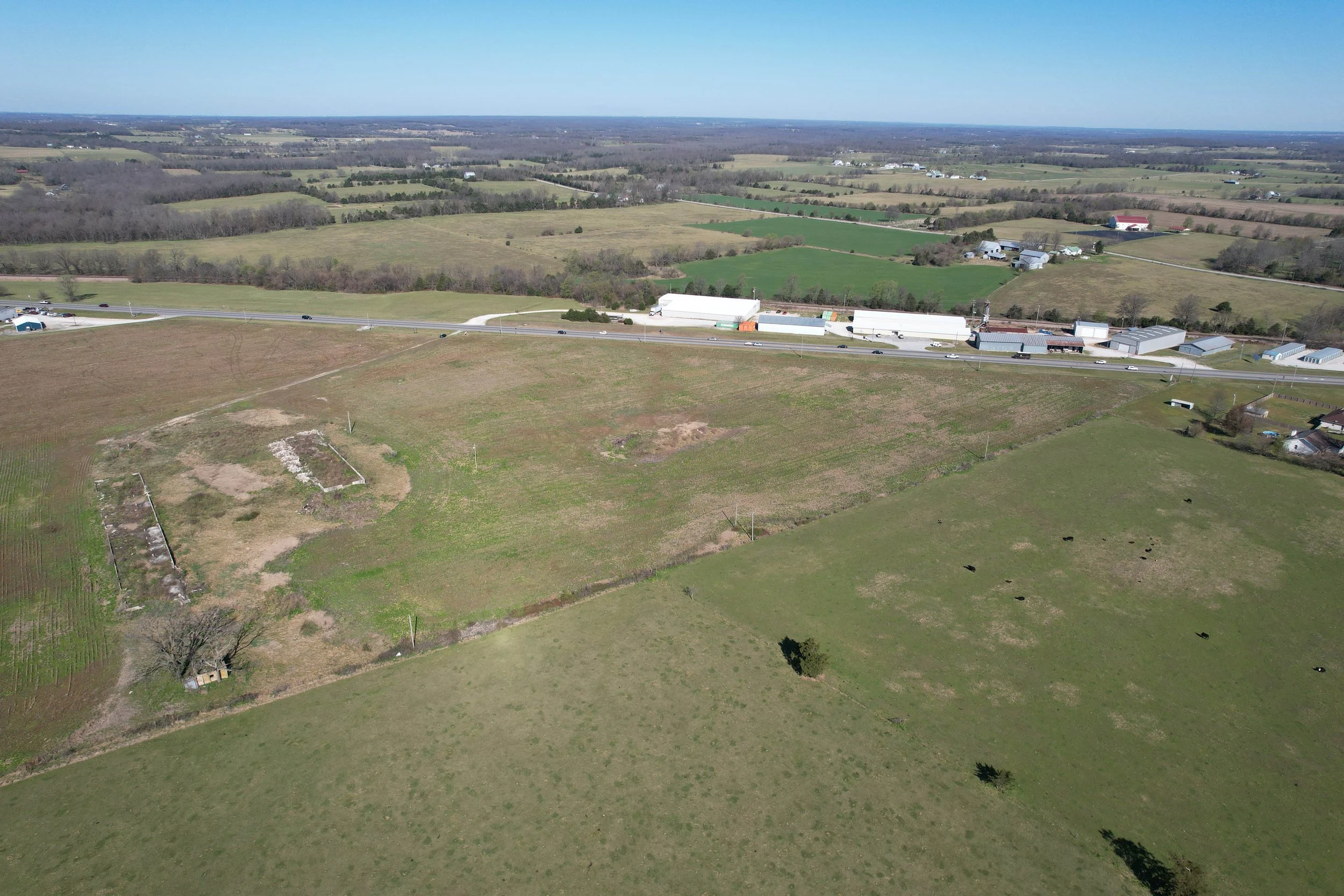 Aerial view of rural farmland with green fields, pastures, scattered houses, trees, and a highway with vehicles, under a clear blue sky.