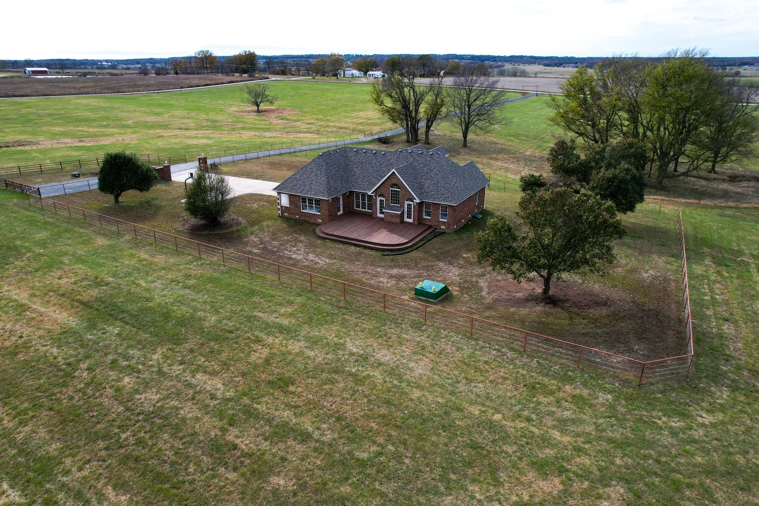 Aerial view of a brick house with a wooden deck, surrounded by trees, on a large grassy farm property with fenced areas and a driveway, with open fields and distant farm buildings in the background.