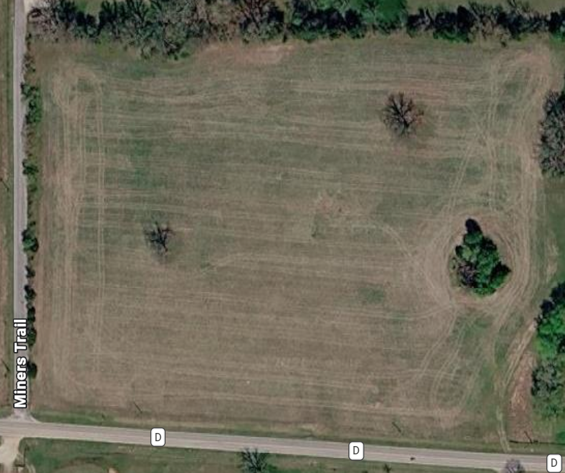 Aerial view of a large grassy field with three small trees and a small pond or water feature, bordered by a road named Miners Trail on the left and another road at the bottom.