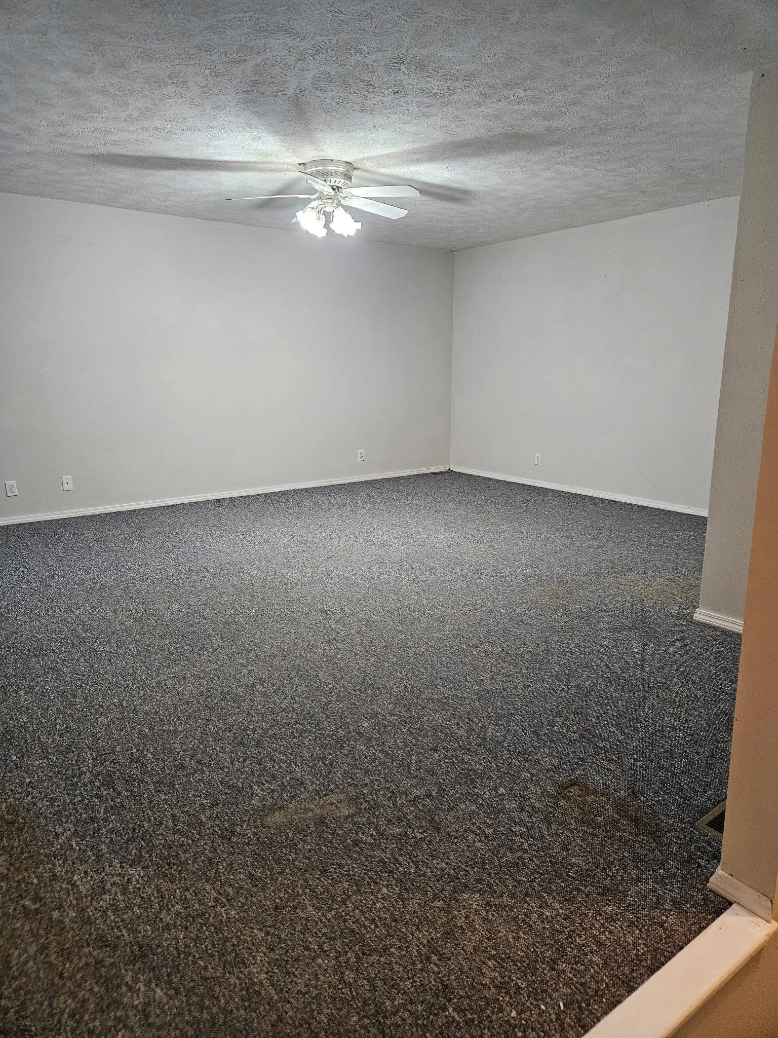 Empty room with gray textured carpet, white walls, a ceiling fan with lights, and a heating vent on the floor.