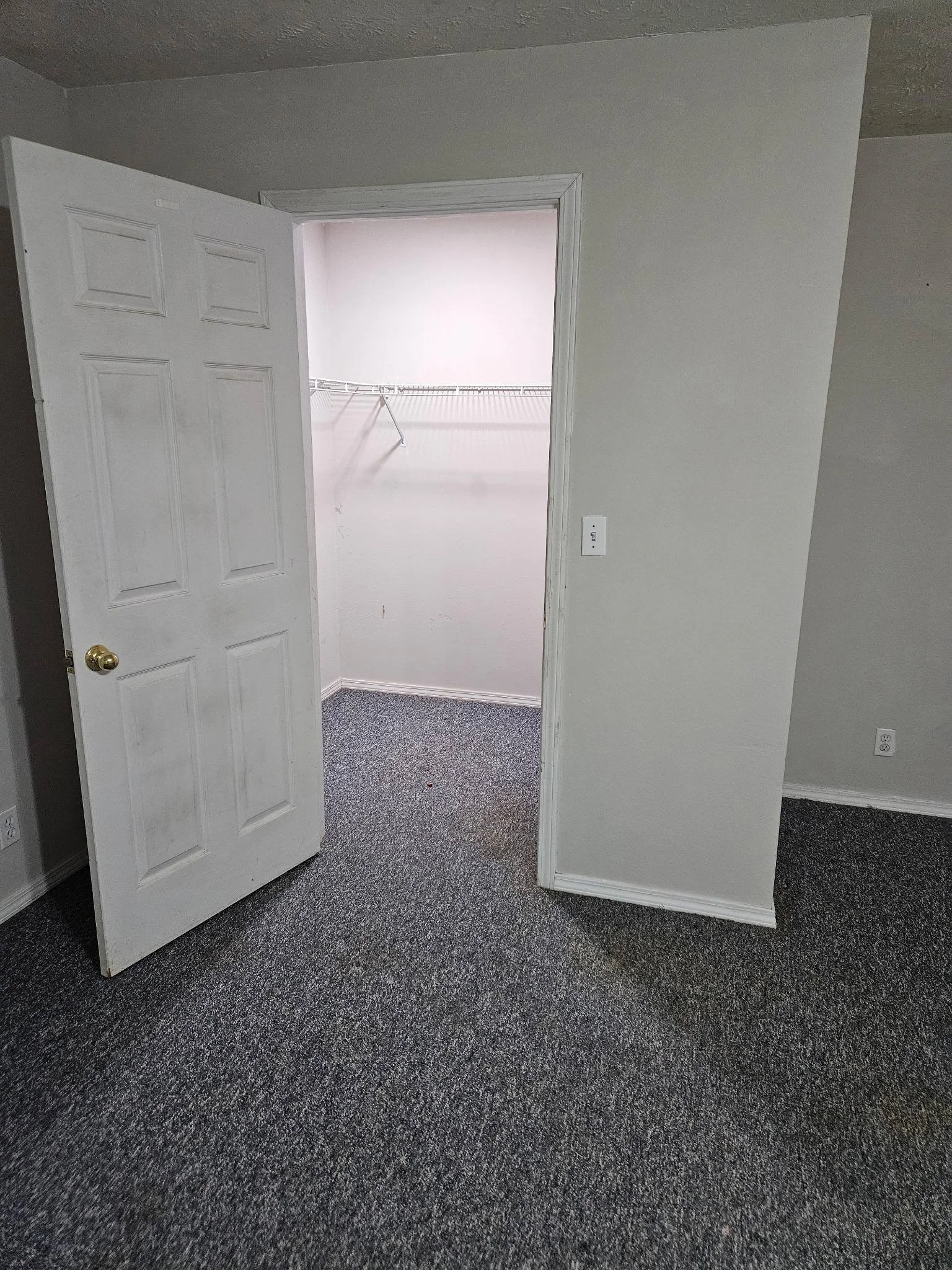 Open closet door revealing an empty walk-in closet with a white wire shelf, in a room with white walls and dark carpeted flooring.