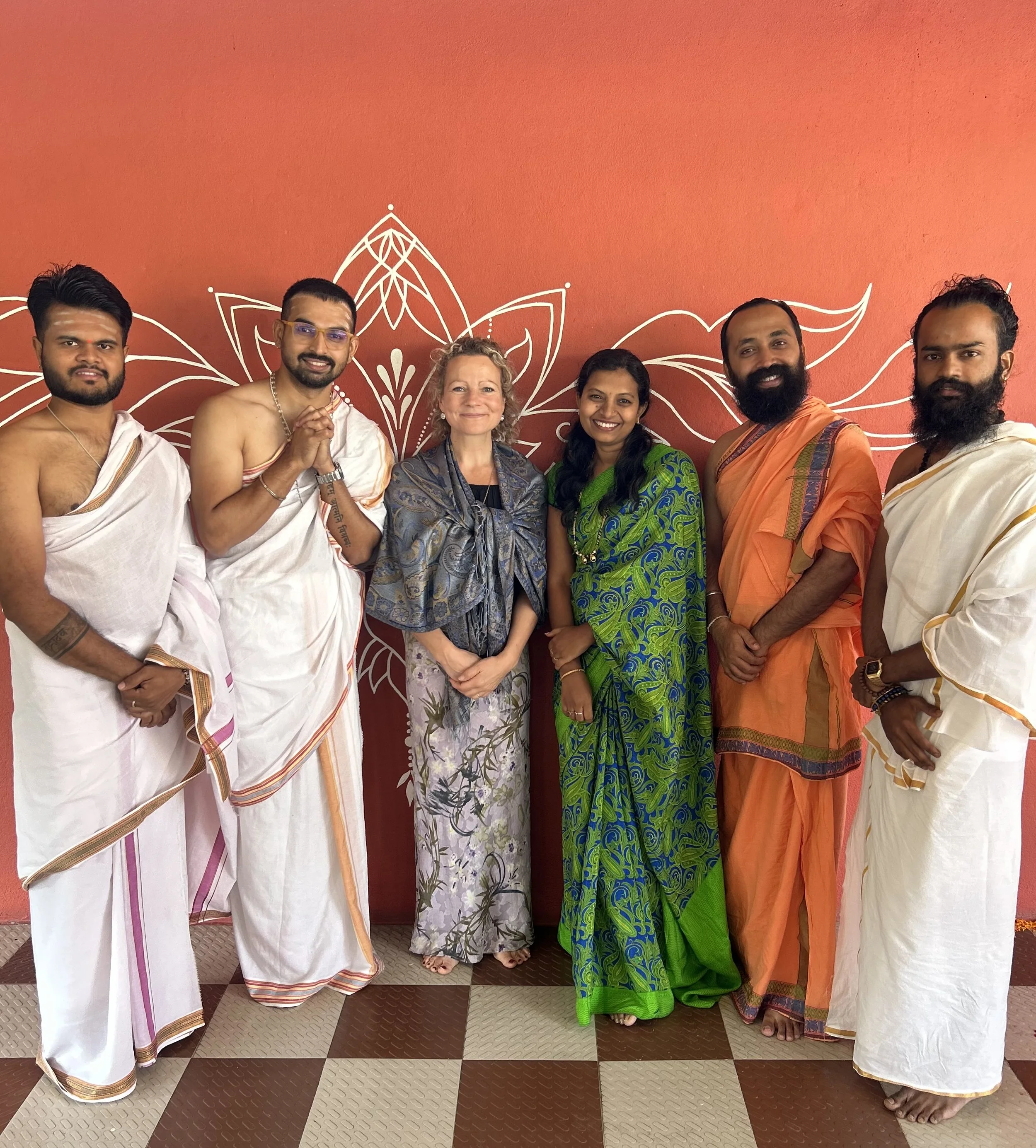 Group of six people standing together indoors in front of a red wall with white decorative art. Four men dressed in traditional Indian attire and two women, one in a green sari and the other in a patterned dress, smiling for the photo.