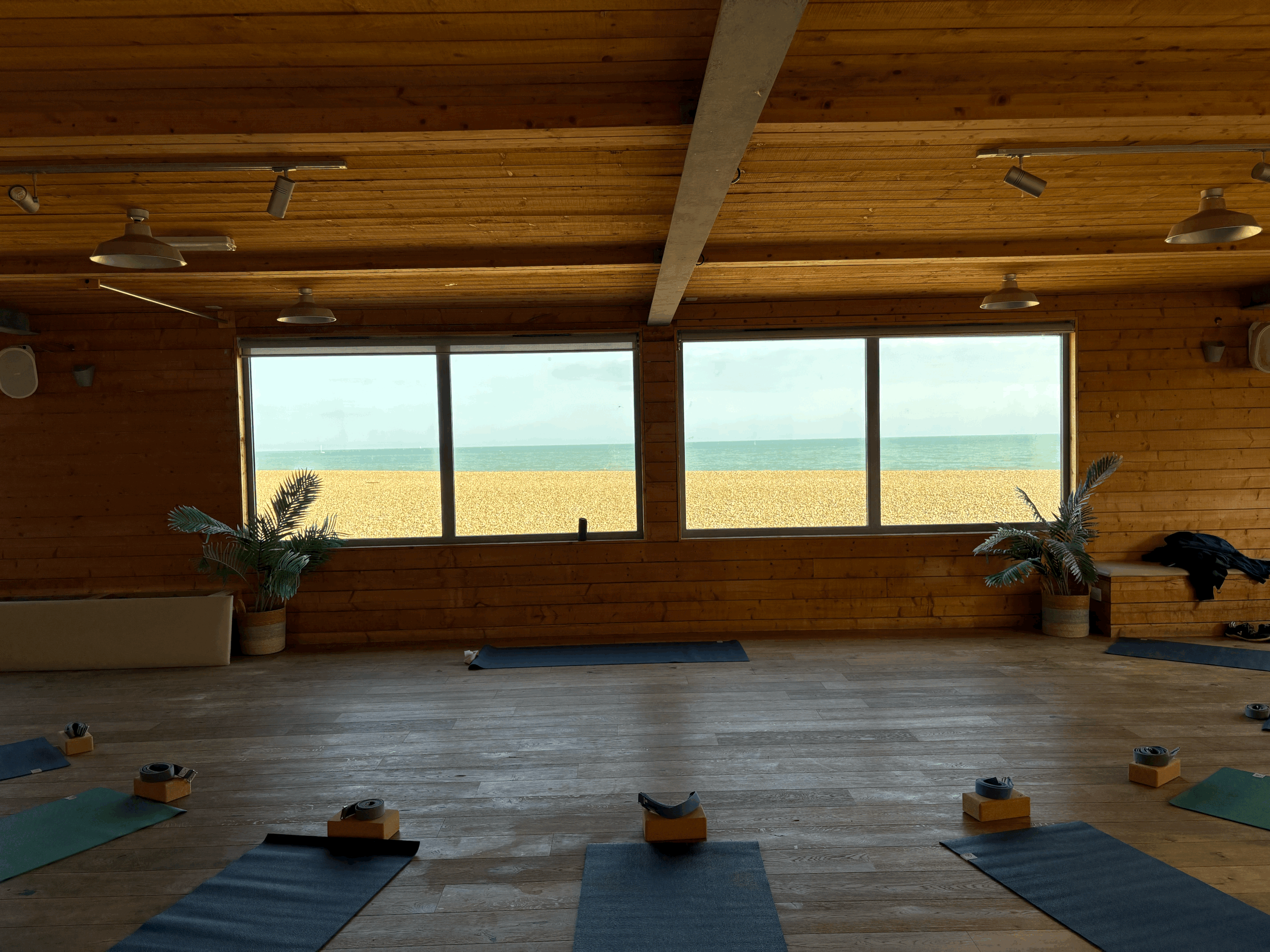 An indoor yoga studio with wooden walls and ceiling, large windows showing a beach and sea view, yoga mats and blocks on the floor, and some plants.