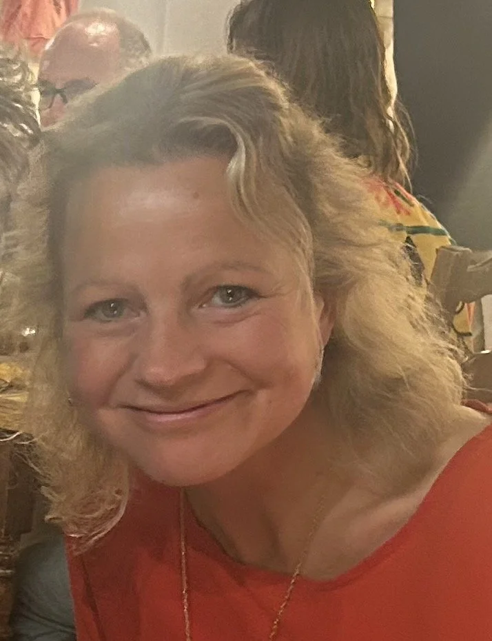 A woman with wavy blonde hair and a red top smiling at the camera in a lively gathering setting.