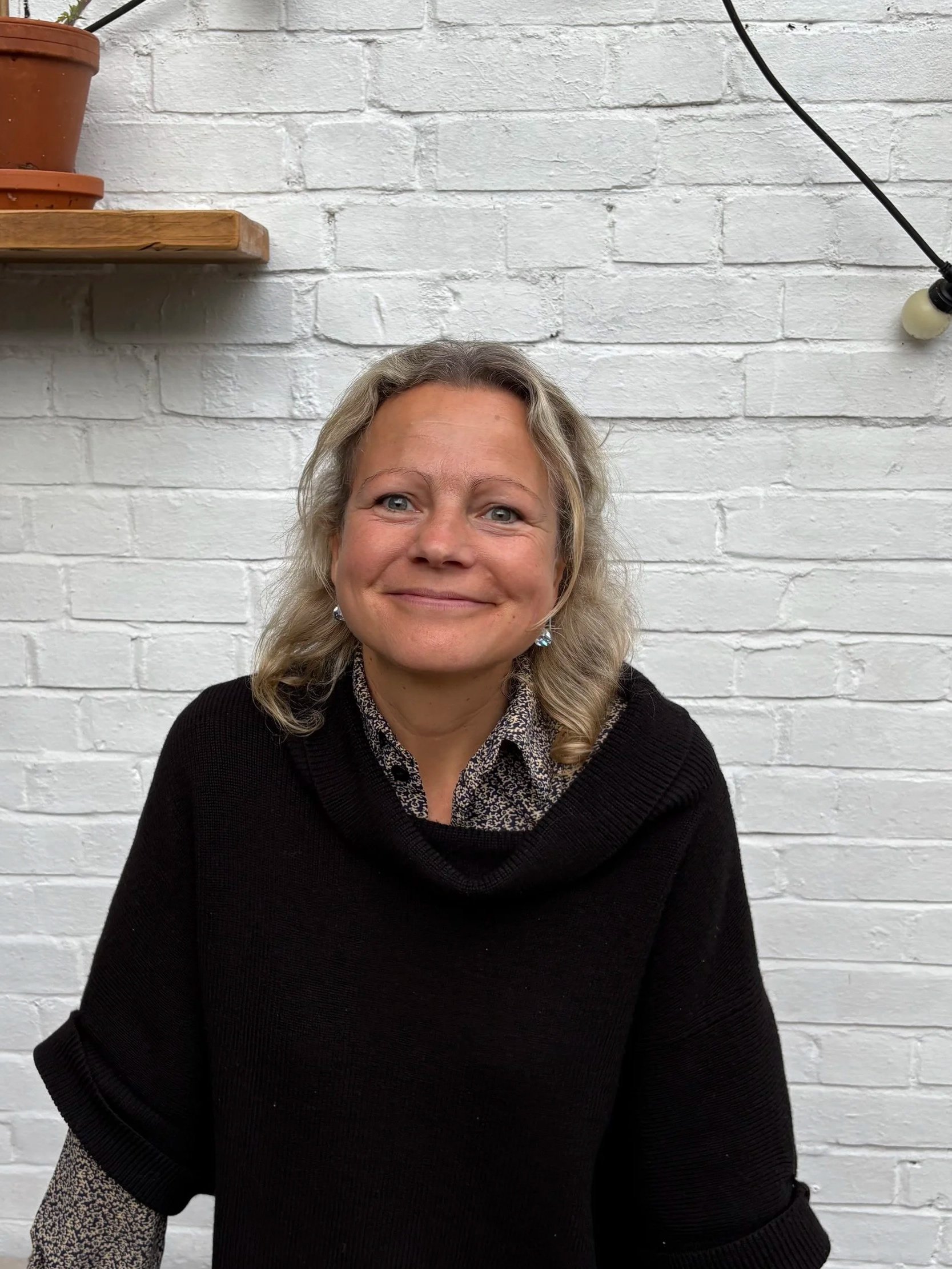 Smiling woman with blonde hair and earrings, sitting in front of a white brick wall, wearing a black sweater and patterned shirt.