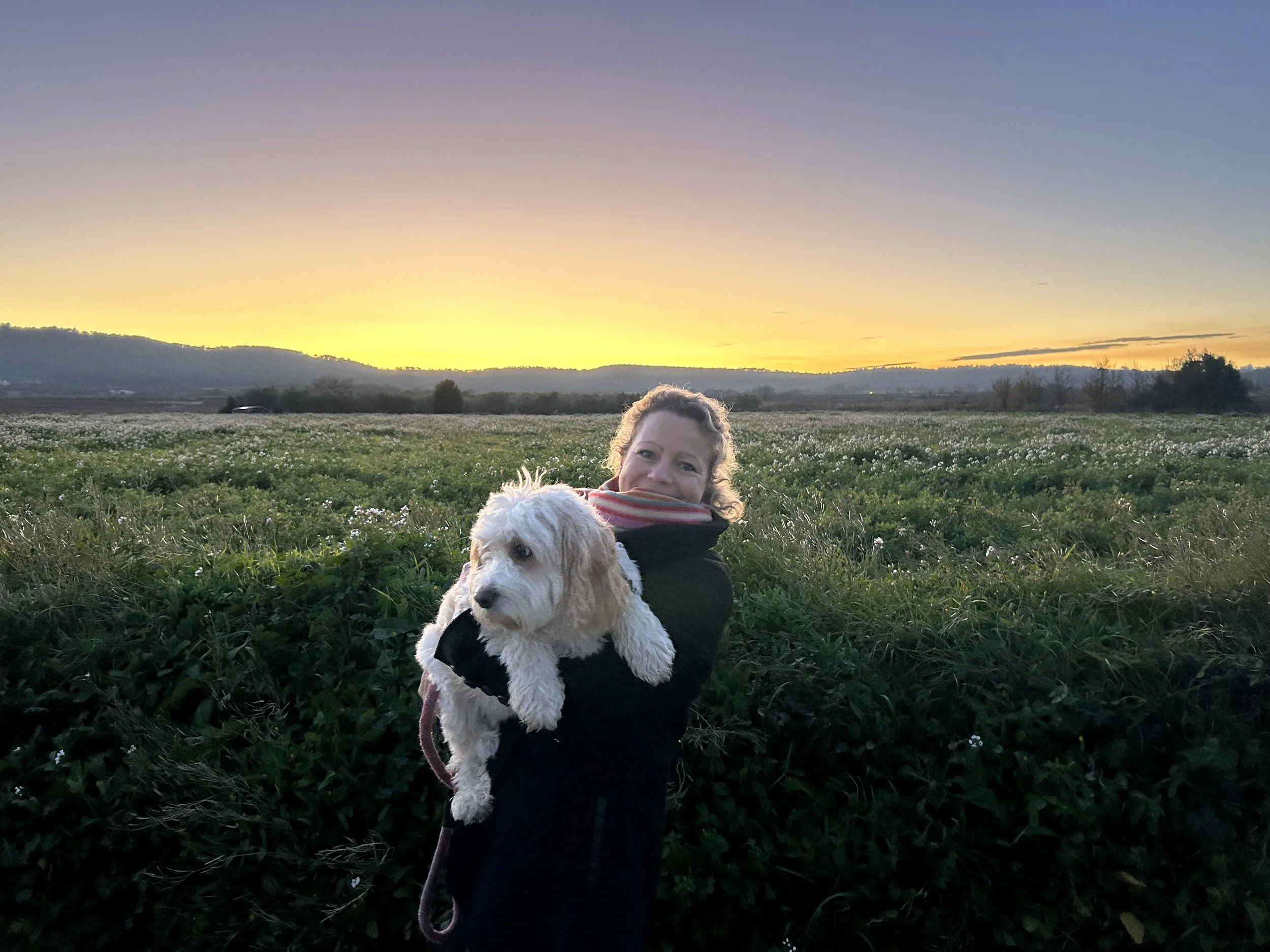 A woman smiling while holding a white dog in a green field at sunset with a mountain range in the background in southern France.