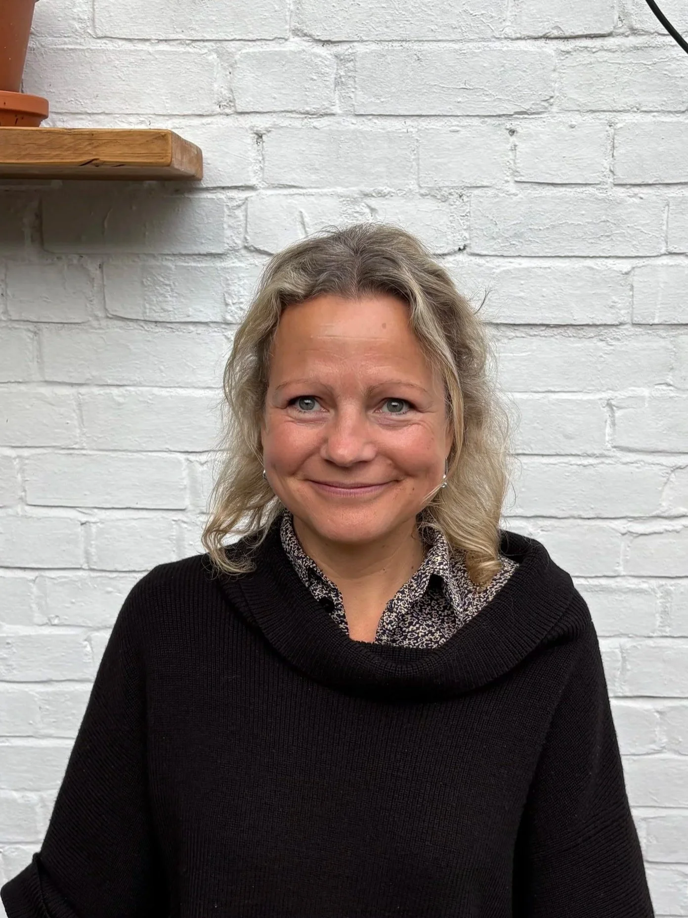 A woman with blonde, curly hair, wearing a black sweater and a patterned shirt, smiling in front of a white brick wall.