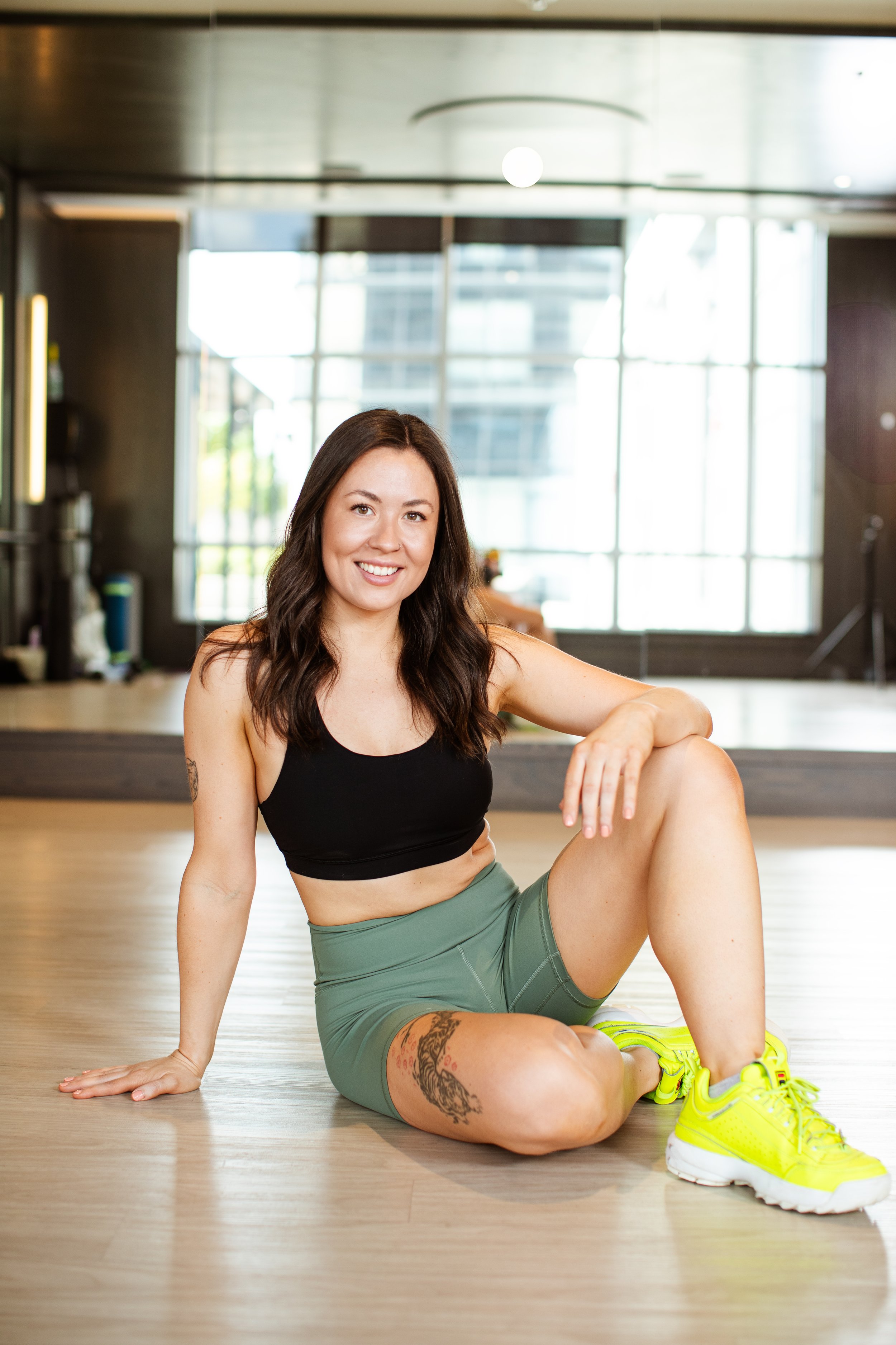 A woman in athletic attire, black sports bra, green shorts, and bright yellow sneakers, sitting on the floor of a gym with large windows and wooden flooring, smiling at the camera.
