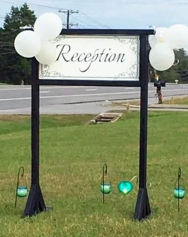 A wedding reception sign mounted on a black frame with white balloons attached, placed on a grassy area near a road.