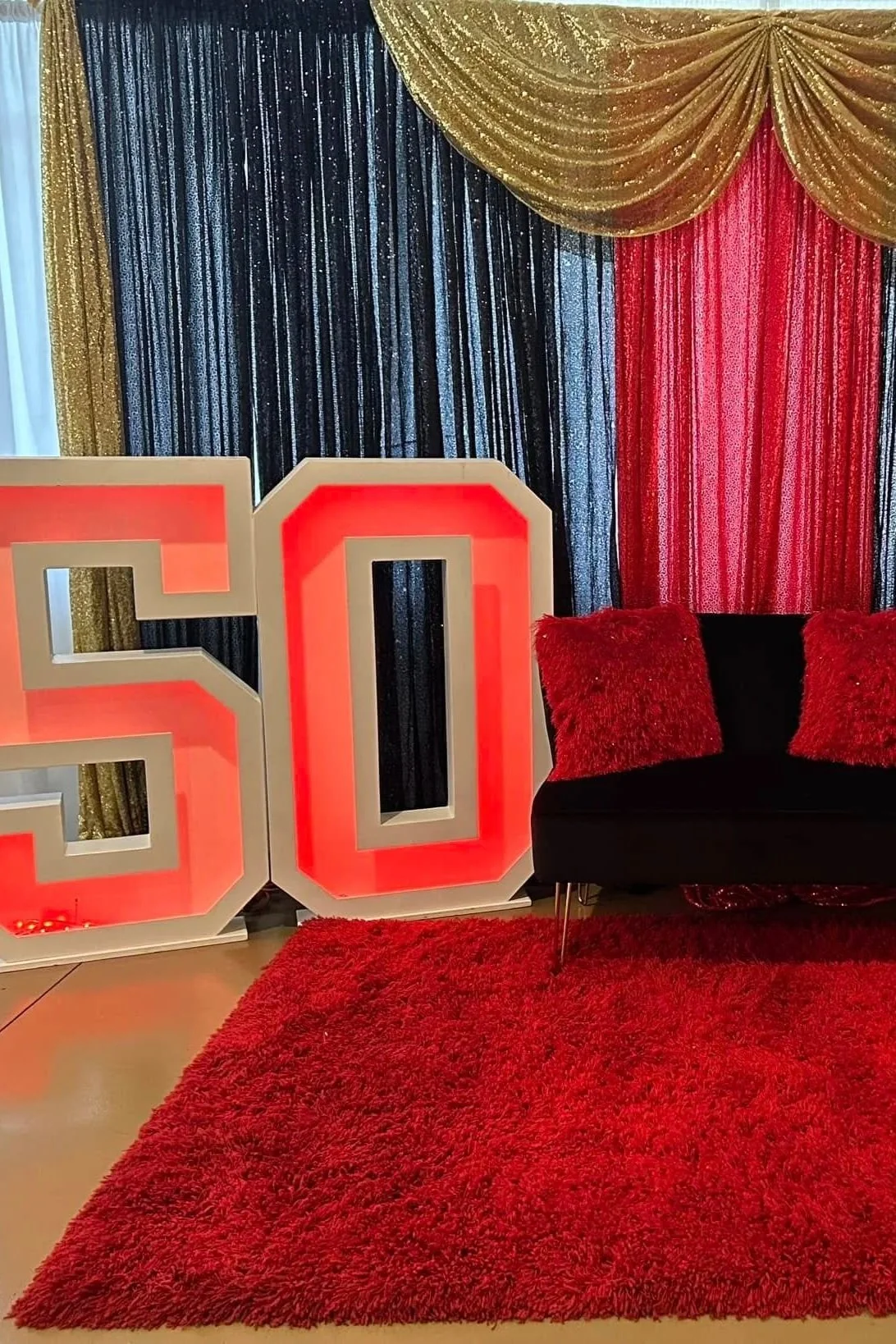 Decorative scene with large illuminated number '50' light in front of a backdrop of gold, black, and red curtains, a black sofa with red pillows, and a red shaggy rug on the floor.