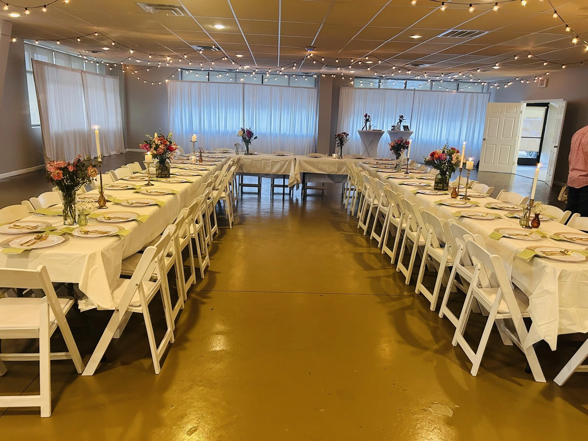 A decorated banquet hall with long white tables covered in white tablecloths, set with plates, napkins, and gold utensils. The tables are adorned with vases of colorful flowers and tall candles in candlesticks. String lights hang from the ceiling, an