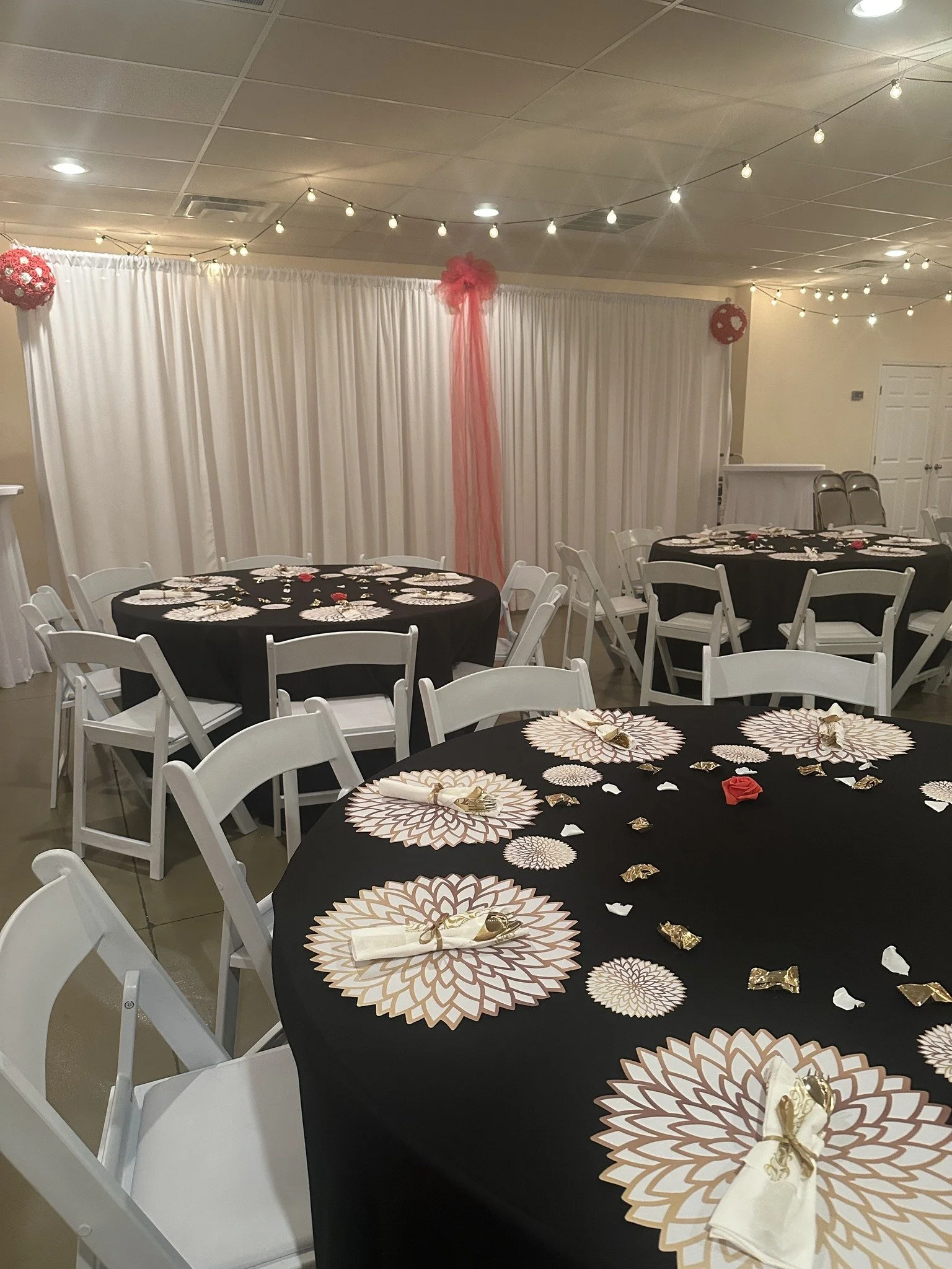 Decorated event room with round tables covered in black tablecloths, white chairs, and floral designs on napkins, set for a celebration or gathering, with string lights and curtain backdrop with pink tulle.