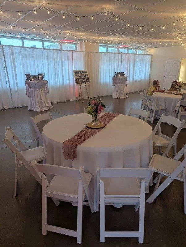 Indoor event space with round tables, white chairs, and string lights on the ceiling, decorated for a celebration.
