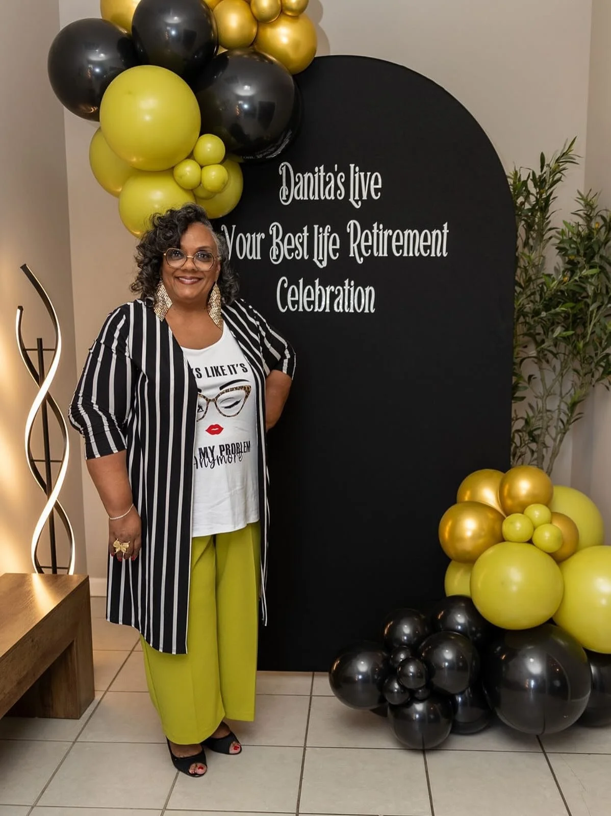 Woman with curly hair and glasses standing next to black and gold balloon decorations, smiling, in front of a black sign that reads 'Janita's live Your Best Life Retirement Celebration,' wearing a striped black and white blazer, a T-shirt with a grap