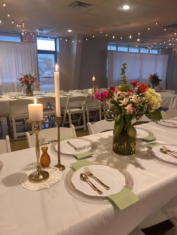 A decorated banquet table set for a formal event with white tablecloths, floral centerpieces, candles, and place settings in a softly lit room with string lights and windows.