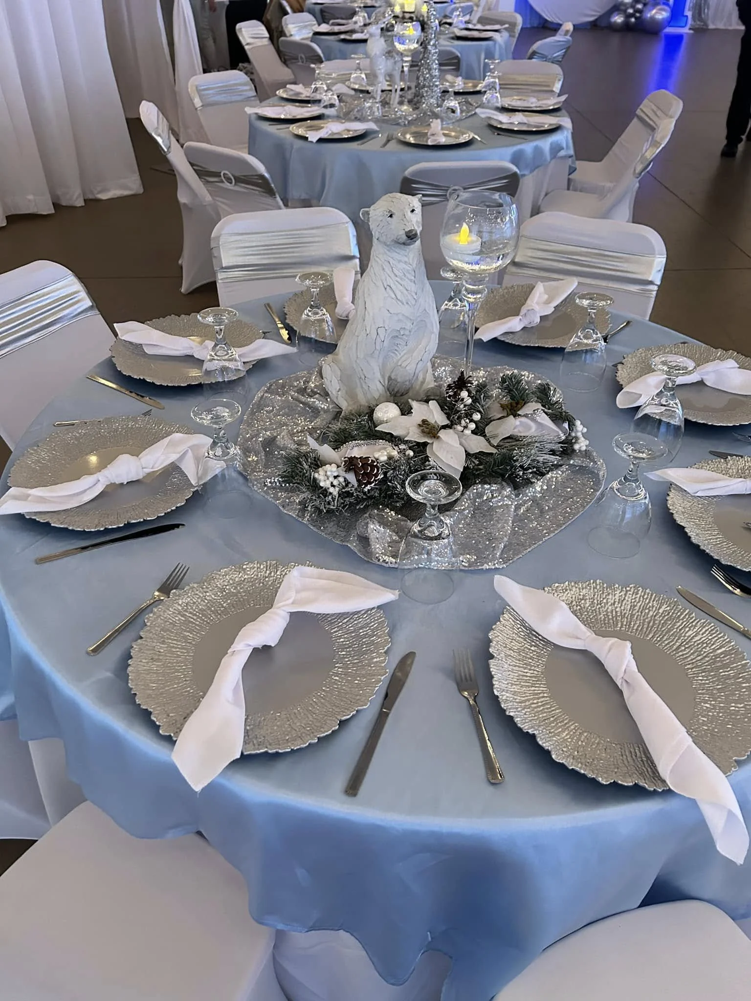 Round table set for a formal event with a wintery centerpiece, white chair covers, blue tablecloth, gold plates, white napkins, and glassware. The centerpiece features a white polar bear sculpture with pinecones, white flowers, and greenery.