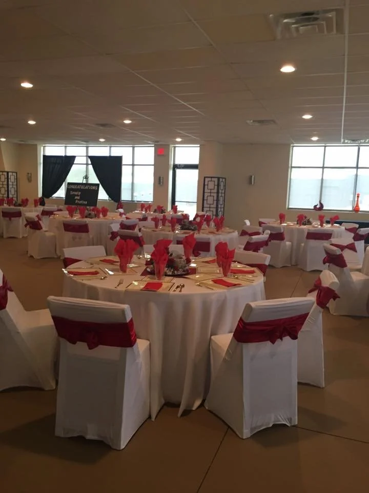 A banquet hall decorated for a celebration with round tables covered in white tablecloths, red napkins, and chairs draped in white with red sash decorations. There is a blackboard in the background with a congratulatory message, and the room has larg