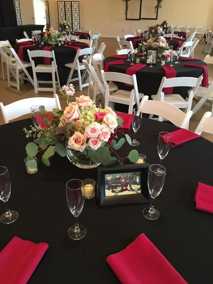 Wedding reception hall with round tables covered in black tablecloths, decorated with pink and white floral centerpieces, pink napkins, and champagne glasses, and white chairs.