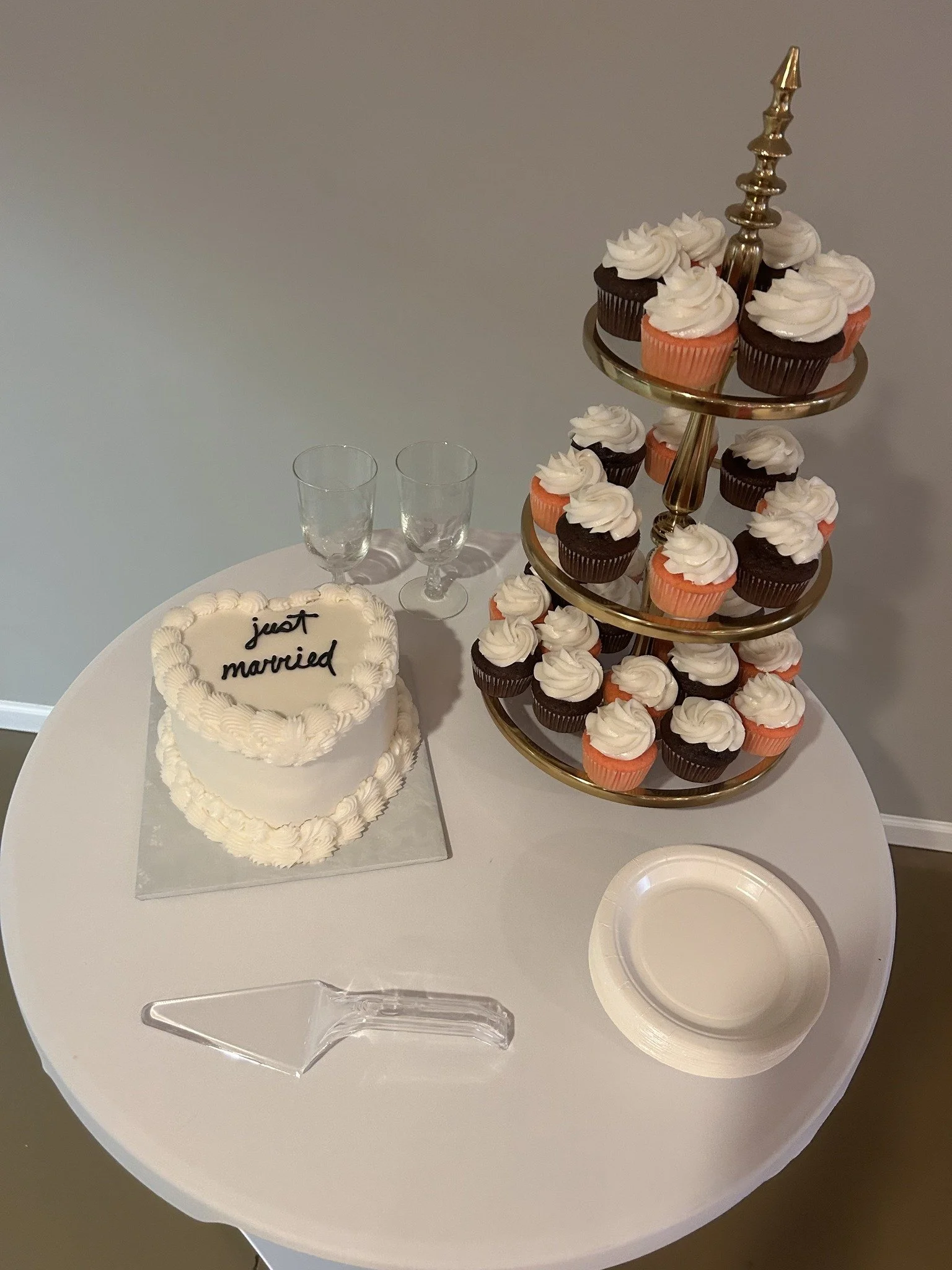 A wedding celebration setup with a heart-shaped cake that has 'just married' written on top, a tiered cupcake stand with chocolate and vanilla cupcakes topped with white frosting, two empty glasses, a paper plate, and a cake server on a white round t