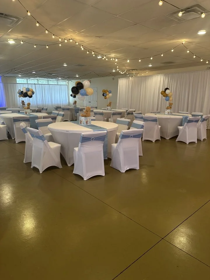 Event hall decorated with string lights, round tables with white tablecloths, and chairs covered with white fabric and blue sashes. Balloons arranged in clusters on the tables and in the room, with teddy bears as centerpieces on the tables. White dra